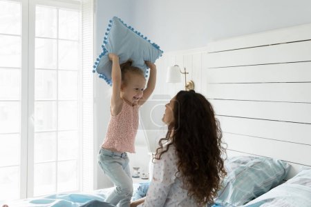 Photo for Joyful little daughter enjoy playtime with young mother in bedroom, girl holding pillow, playing with mommy, fighting together in morning at home. Happy active family leisure, games with kids, ties - Royalty Free Image