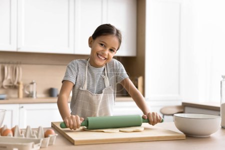 Photo for Happy beautiful baker kid girl in household apron preparing bakery meal in kitchen, cooking pastry dessert for family dinner, rolling raw dough on table, looking at camera, smiling for portrait - Royalty Free Image