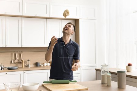 Photo for Cheerful young baker man throwing raw dough up, having fun while cooking at home, standing at kitchen table with roller, flour, furniture and utensil in background, laughing, enjoying culinary - Royalty Free Image