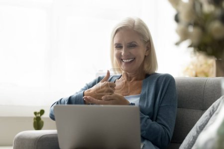 Smiling mature woman with hearing impairment disorder sit on sofa with laptop, take part in virtual meeting, engaged in inclusive remote deaf community communication using sign language at video call