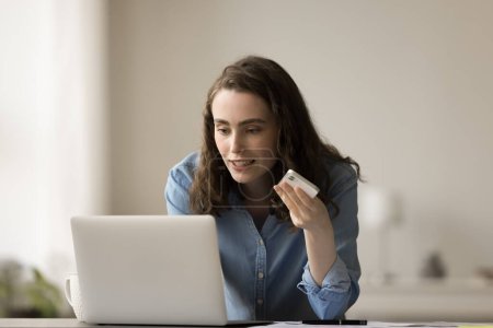 Positive freelance worker girl speaking at mobile phone, holding gadget at face, recording voice message, listening to audio file, working at laptop at home office workplace table