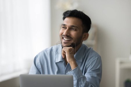 Téléchargez les photos : Heureux jeune homme d'affaires indien professionnel pensant à la table de travail à la maison, regardant la fenêtre, souriant, riant, travaillant à l'ordinateur portable, planifiant le projet réussi, succès de carrière - en image libre de droit