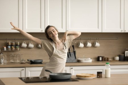 Positive carefree baker girl in apron dancing at table with bakery food, cooked crepes, dish, enjoying motion, music, cooking hobby, culinary, preparing breakfast, smiling, laughing