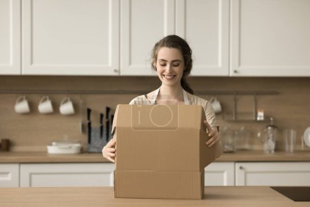Photo for Happy beautiful young customer woman opening package, unpacking cardboard box, parcel on kitchen table, looking inside, smiling, getting delivered purchase from Internet shop, grocery store - Royalty Free Image