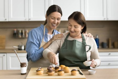 Téléchargez les photos : Confiseur positif joyeux maman et jeune fille adulte femme décorer cupcakes au chocolat maison, préparer le dessert savoureux sucré pour la fête de famille ensemble - en image libre de droit