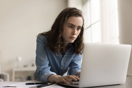 Téléchargez les photos : Femme d'affaires sérieuse jeune génération Z tapant à l'ordinateur portable, se penchant sur la table de travail engagée dans le processus, regardant l'affichage, écrivant idée créative pour le projet - en image libre de droit