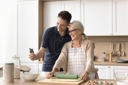 Photo for Happy young adult man and senior blonde mom baking in kitchen and talking on video call on smartphone, holding mobile phone, hugging, looking at gadget screen, smiling, laughing - Royalty Free Image