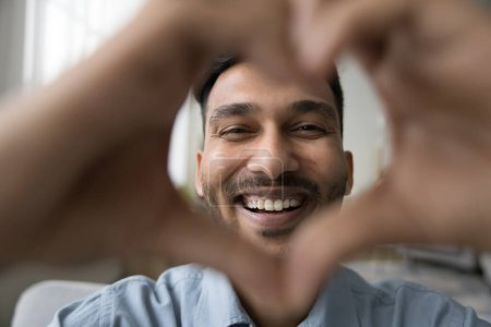 Photo for Cheerful attractive young Indian man smiling at camera through hand heart frame. Happy joyful attractive guy showing romantic symbol of love, Valentines day. Indoor male portrait - Royalty Free Image