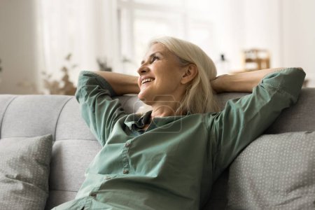 Foto de Mujer mayor rubia atractiva feliz relajándose en cómodo sofá acogedor, apoyado en la espalda suave, soñando despierto, mirando hacia otro lado, sonriendo, pensando en una buena jubilación, disfrutando de la comodidad - Imagen libre de derechos