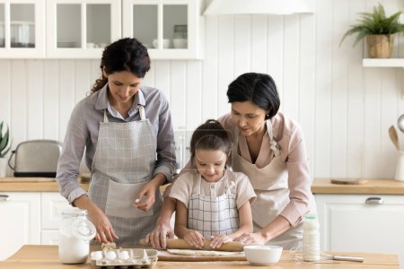 Zwei Mehrgenerationenfrauen mit Kind kochen gemeinsam in der gemütlichen, häuslichen Küche. Junge Frau ihre kleine Tochter und reife Mutter tragen Schürzen und bereiten am Wochenende zu Hause selbst gebackenes Gebäck zu