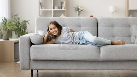 Photo for Happy positive lazy pre teen kid girl enjoying leisure, relaxation, comfort at home, resting on comfortable sofa, lying on side, looking at camera, smiling, laughing. Full length portrait - Royalty Free Image