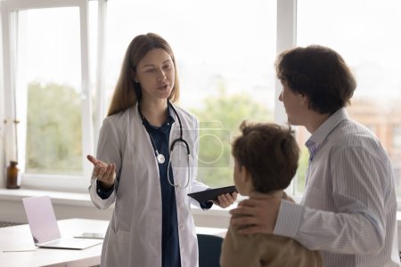 Photo for Positive pediatrician in white coat with stethoscope meeting with little patient kid and parent, talking to father, giving medical consultation after examining boy in doctors office - Royalty Free Image