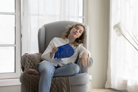 Tired relaxed beautiful young woman suffering from heat, hypoxia, stuffy air, cooling hot air, waving paper handheld fan with closed eyes, feeling relief, resting in armchair at home