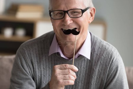 Téléchargez les photos : Heureux homme âgé positif appliquant la fausse moustache de papier sur le bâton à face, célébrant movember, regardant l'appareil photo, s'amusant, souriant, riant, en utilisant un accessoire de tir photo. Portrait recadré - en image libre de droit