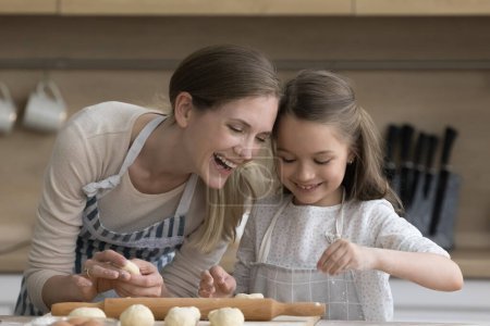 Téléchargez les photos : Joyeux jolie mère et heureuse petite fille enfant dans des tabliers cuisiner ensemble, façonner, pétrir la pâte, préparer des tartes, biscuits, sourire, rire, profiter de l'activité culinaire à la maison - en image libre de droit