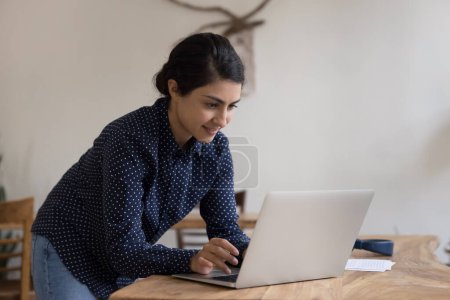 Photo for Focused happy young Indian laptop user woman standing at workplace table in stylish home office, typing, using online app, enjoying wireless Internet communication with client - Royalty Free Image