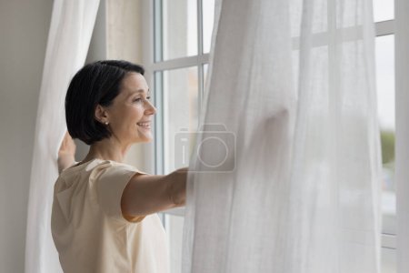 Happy mature homeowner woman looking out of big window, enjoying view, daylight, good morning, parting white transparent veils in bedroom, opening drapes, curtains