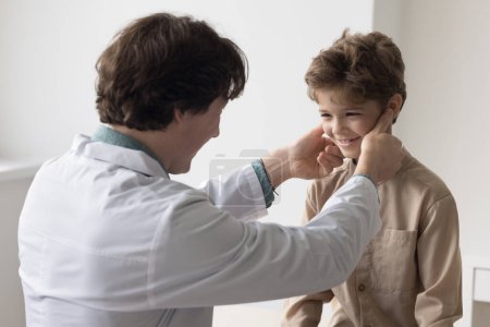 Foto de Amistoso pediatra masculino examinando niño, tocando los ganglios linfáticos debajo de las mandíbulas. Médico chequeando sonriente chico positivo para infección respiratoria, virus, gripe en la clínica - Imagen libre de derechos