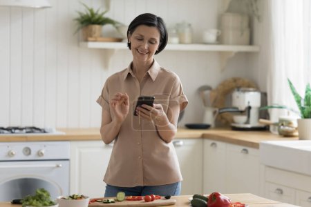 Téléchargez les photos : Joyeux heureux femme chef mature utilisant un téléphone mobile sur la table avec des ingrédients alimentaires biologiques, salade de cuisine, lecture de la recette sur Internet, prendre des photos, sourire, rire - en image libre de droit