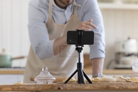 Photo for Male baker preparing pastry food, speaking at smartphone webcam placed on kitchen table. Cook blogger recording video workshop of baking pies. Close up of mobile phone fixed on holder - Royalty Free Image