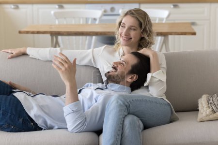 Photo for Happy relaxed young couple in love resting on sofa at home, hugging, sitting, lying on couch, looking away, talking, smiling, laughing, discussing plans, enjoying romantic relationship, leisure - Royalty Free Image