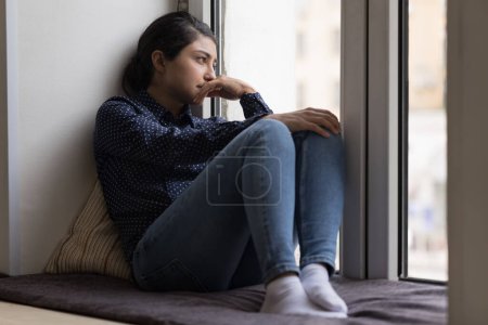 Téléchargez les photos : Dépression contrariée Femme indienne reposant sur un rebord de fenêtre souple, regardant par la fenêtre perdue dans de mauvaises pensées négatives, traversant une crise émotionnelle, dépression, stress, tristesse, frustration - en image libre de droit