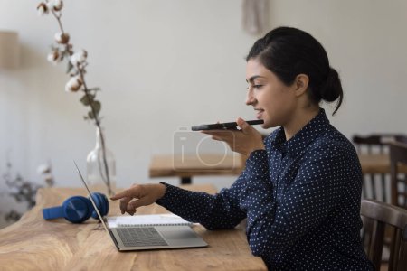 Positive young freelance Indian business woman using app on gadgets for online communication, recording voice message on smartphone, pointing at laptop computer monitor, working at desk in home office