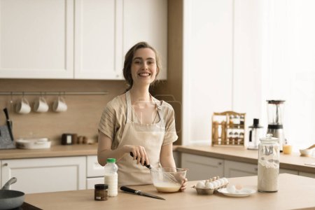 Cheerful pretty adult baker girl in apron enjoying culinary hobby in kitchen, preparing dough for pancakes, cooking omelet, beating raw eggs in bowl, looking at camera, smiling for portrait