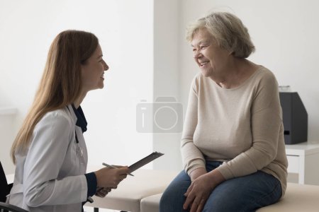 Photo for Elderly 80s patient woman visiting practitioner for examination, sitting on medical couch, talking to positive doctor woman, smiling, laughing. Physician meeting with old lady, giving recommendations - Royalty Free Image
