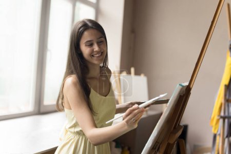Photo for Happy inspired art school student girl enjoying creative hobby at easel, drawing picture in acrylic paints, applying white paint with brush, holding palette, smiling, laughing - Royalty Free Image