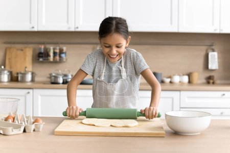 Photo for Happy preteen baker child girl in chef apron baking alone in home kitchen, rolling dough for pitta on table with bakery food ingredients, preparing homemade pastry dessert, enjoying culinary hobby - Royalty Free Image