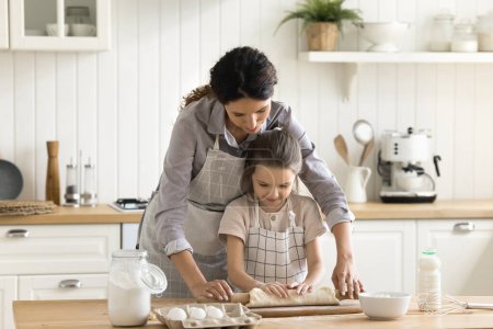 Jeune mère hispanique aimante enseigner à sa fille préscolaire à cuisiner, aplatir la pâte pour les pâtisseries faites maison, profiter du temps de cuisson dans une cuisine confortable. Activité du week-end familial, partager les connaissances avec la progéniture