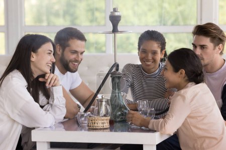 Multiracial group of happy cheerful young friends smoking hookah in lounge cafe, sitting at table, drinking tea, talking, chatting, laughing, enjoying meeting, leisure time, break