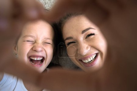 Photo for Happy excited Asian little kid girl and young mom showing hand heart, looking at camera through finger frame, smiling, laughing, having fun, lying on floor, posing for close up shot - Royalty Free Image