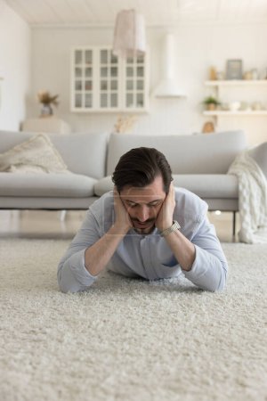 Photo for Tired sad millennial Caucasian man feeling depressed, suffering from headache, lying on carpeted floor at home, touching head, feeling apathy, boredom, fatigue, burnout - Royalty Free Image