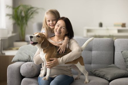 Photo for Cheerful grandmother and dog owner woman cuddling beagle, piggybacking girl on soft couch, laughing, smiling for portrait, looking at camera, relaxing at home with granddaughter and beloved pet - Royalty Free Image