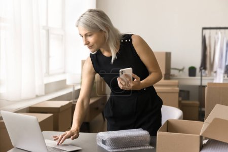 Photo for Busy senior entrepreneur woman using gadgets for processing Internet store order, sending product to client, packing cardboard box at home storage place, holding smartphone, typing on laptop - Royalty Free Image