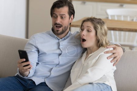 Foto de Impresionante pareja joven confundida recibiendo inesperadas noticias sorprendentes, mirando el teléfono inteligente, leyendo el mensaje de texto, aviso, jadeando, sentado en el sofá en casa, abrazándose - Imagen libre de derechos