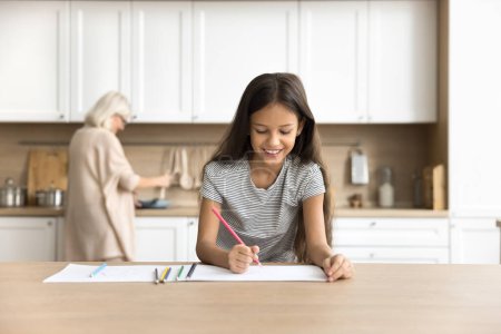 Photo for Happy preteen kid girl drawing in colorful pencils in home kitchen while blonde grandma cooking lunch in blurred background. Positive schoolchild doing school homework task at home - Royalty Free Image