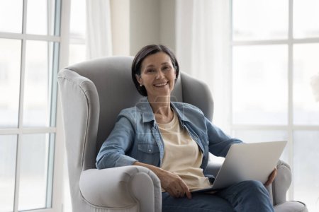 Photo for Happy pretty mature freelance professional woman home portrait. Cheerful business lady using laptop for remote job, relaxing in armchair, looking at camera, smiling - Royalty Free Image