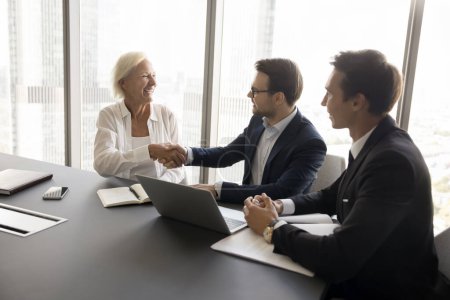 Photo for Happy mature business woman giving handshake to colleague man at meeting table. Younger boss thanking mature professional for good job, successful idea, shaking hand, smiling - Royalty Free Image