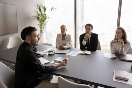 Photo for Confident male professional talking to coworkers on corporate meeting, sitting at conference table, sharing ideas for brainstorming. Candidate speaking to HR team on job interview - Royalty Free Image