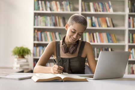 Photo for Positive student girl studying in campus library, writing notes, listening to webinar, online lesson on laptop, doing academic research study, enjoying education, knowledge - Royalty Free Image