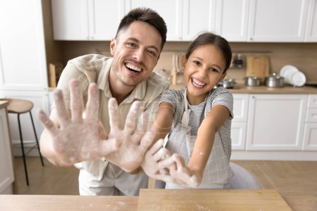 Cheerful baker dad and laughing daughter child having fun together, making mess, making together in home kitchen, showing floury hands, looking at camera, smiling for portrait