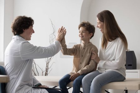 Foto de Positive cheerful doctor man giving high five to patient kid. Happy pediatrician meeting with boy and mom in clinic office, discussing checkup good result, clapping hands with healthy child - Imagen libre de derechos