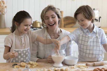 Joyful young mother and two happy pretty little kids in aprons baking together in home kitchen, having fun, laughing at floury smudges on faces, shaping dumplings, cookies from dough at table