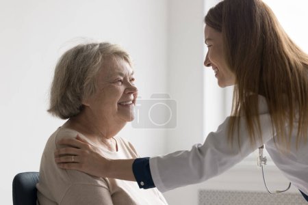 Photo for Happy pretty doctor woman giving comfort to positive older senior patient, touching shoulder, speaking, smiling, laughing. Elder lady visiting practitioner in office - Royalty Free Image