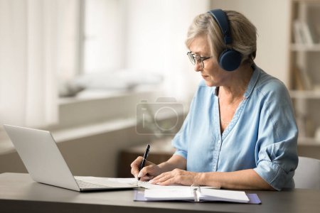 Photo for Focused senior woman in glasses and earphones working at laptop computer at home, watching training educational lecture, course, writing notes, getting new occupation, profession - Royalty Free Image