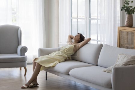 Peaceful calm sleepy young adult girl enjoying relaxation at cozy stylish home with pale interior colors, resting on soft couch with hands on nap, sleeping, taking deep breath of fresh air
