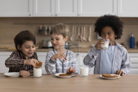 Diverse team of little kids drinking milk in home kitchen, eating cookies, chatting, laughing, having fun, enjoying snacks. Happy boys keeping healthy nutrition, getting calcium from dairy products